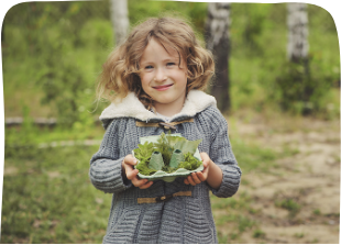 Smiling child outdoors holding a plate of fresh green leaves wearing a cozy coat in a natural forest setting