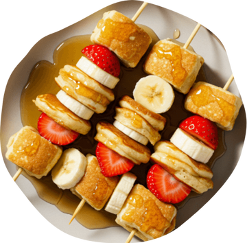 Three skewers with mini pancake pieces, banana slices, and strawberries are arranged on a light-colored plate, drizzled with natural maple syrup—a fun and tasty treat perfect for kids.