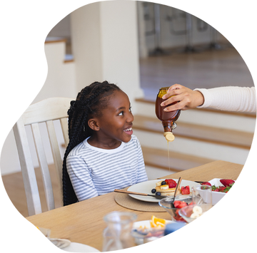 Child smiling at syrup being poured on breakfast with berries and fruit on the table promoting maple-powered natural kids snacks