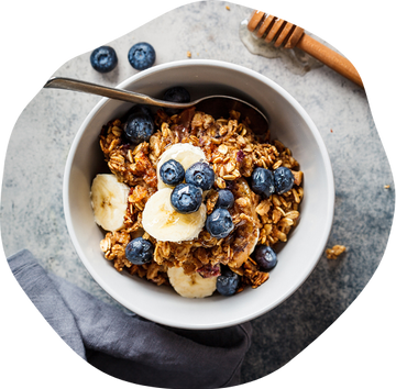 Bowl of granola with banana slices and blueberries next to honey dipper and blueberries on a stone surface
